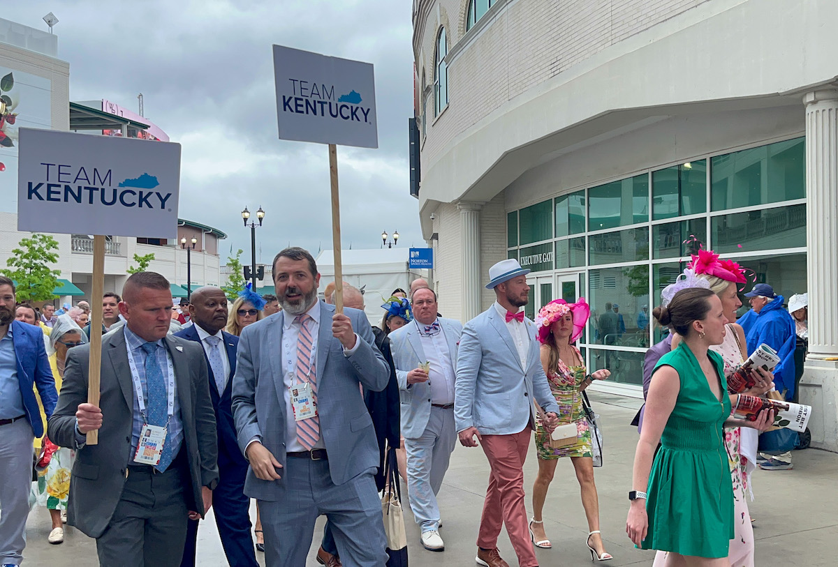 A train of people following Team Kentucky signs walk pass the paddock area of Churchill Downs. Team Kentucky is a Beshear administration slogan. (Kentucky Lantern photo by McKenna Horsley)